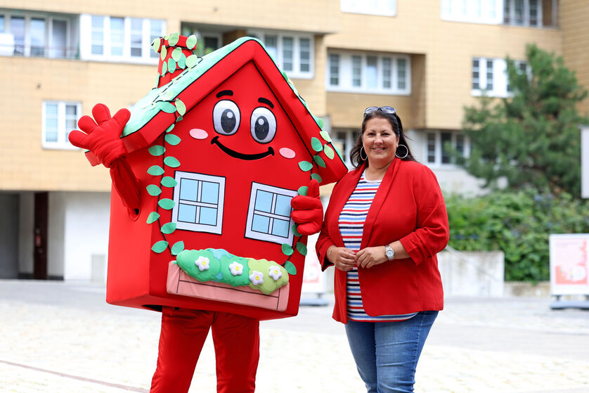 Vizebürgermeisterin und Wohnbaustadträtin Kathrin Gaál posiert neben dem Maskottchen für den Wiener Wohnbau, der Wohna Lisa. Das Kostüm der Wohna Lisa besteht aus einem roten Haus mit viel Blumenschmuck an den Fenstern, Augen, Mund, Armen und Beinen.