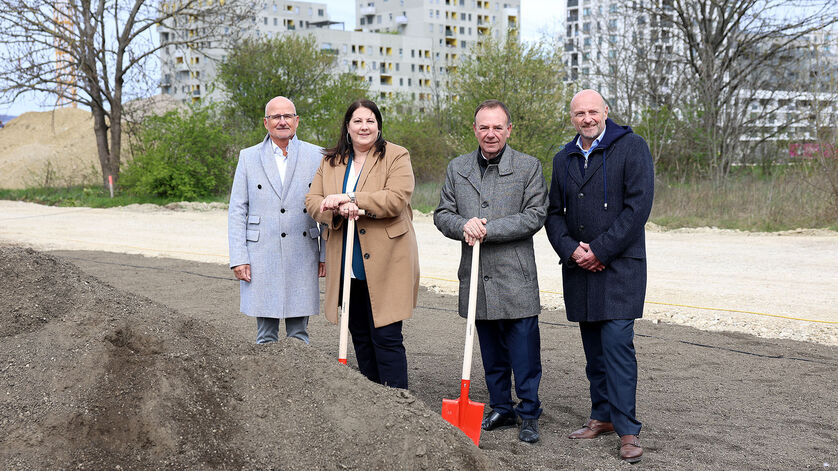 Dieter Groschopf, stellvertretender Geschäftsführer des wohnfonds_wien, Vizebürgermeisterin und Wohnbaustadträtin Kathrin Gaál, Bezirksvorsteher Gerald Bischof und WIGEBA-Vorstand Paul Steurer stehen an Spaten lehnend bei einem Sandhaufen. Die Aufnahme wurden offenbar zu einer kälteren Jahreszeit gemacht, da alle Protagonisten mit warmer Kleidung zu sehen sind.