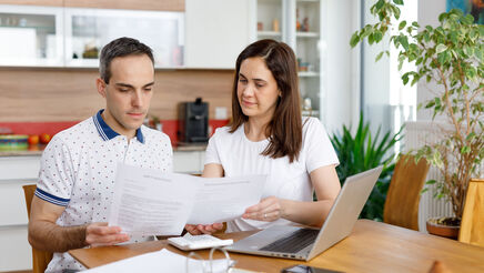 2 Personen sitzen an einem Holztisch und haben ein Notebook vor sich stehen. Zusätzlich hantieren sie mit einigen Zetteln und schauen diese konzentriert an. Im Hintergrund ist eine Küche erkennbar.