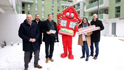5 Personen stehen in winterlicher Business-Bekleidung im verschneiten Innenhof einer Neubauanlage. Ihnen zur Seite wurde wurde Wohna Lisa, das Maskottchen des geförderten Wohnbaus in Wien. Es ist eine als Haus verleidete rote Figur in Form eines Hauses mit Satteldach, großen Augen und einem freundlichen Lächeln.