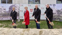 4 Personen stehen mit Spaten mit Sand darauf hinter einem Sandhaufen und lächeln in die Kamera. I Hintergrund ist ein Bauzaun mit Transparenten auf denen Visualisierungen des Gemeindebau NEU und das Logo von Stadt Wien/Wiener Wohnen zu erkennen ist.