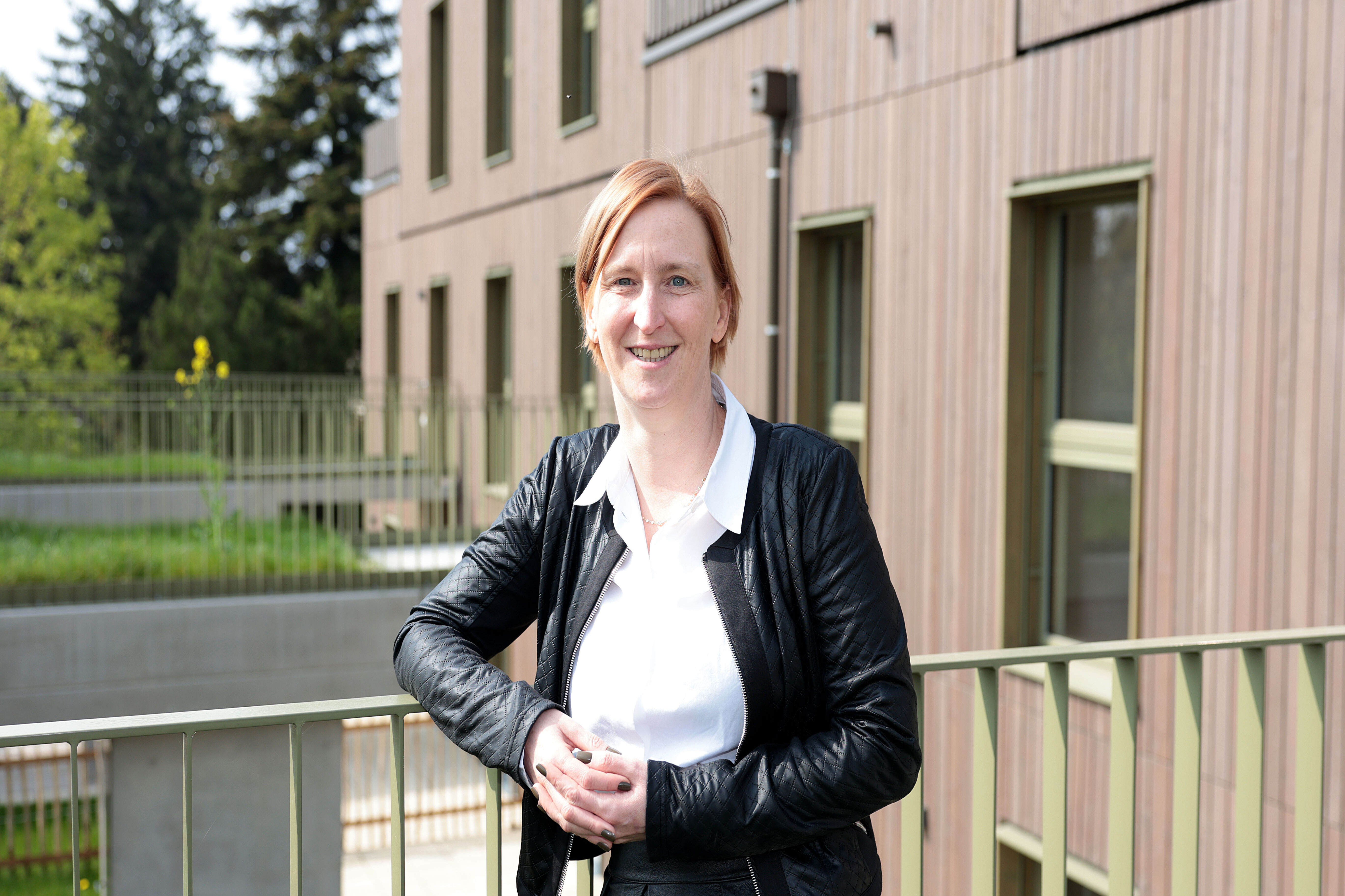 Frau mit schwarzer Lederjacke und weißem Hemd lehnt an einem Geländer vor einem modernen Wohngebäude mit Holzfassade