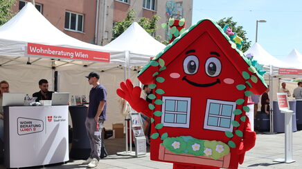 Das wienwohntbesser-Maskottchen Wohna Lisa, ein rotes Haus mit Armen und Beinen, einem Satteldach, einem freundlichen Lächeln im Gesicht und mit ausreichend Blumenschmuck steht auf einem Platz und winkt in die Kamera. Im Hintergrund ist ein Pagodenzelt sichtbar in dem an einem Pult mit Laptops darauf, zwei Personen eine weitere Person beraten, die davor steht.