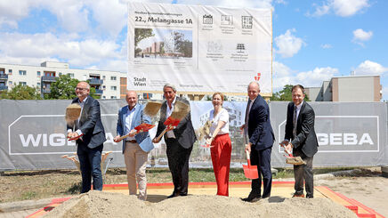 Sechs Personen mit Spaten stehen auf einer Baustelle vor einem großen Schild und schaufeln Erde in eine Sandkiste
