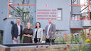 Vier Personen stehen vor einem modernen Wohnhaus mit der Aufschrift 'Wohnhaus der Gemeinde Wien errichtet in den Jahren 2023-2026'