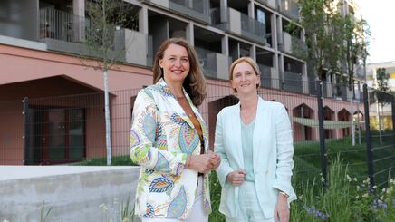 Zwei Frauen stehen vor einem modernen Wohngebäude, eine trägt einen gemusterten Blazer, die andere einen hellen Anzug