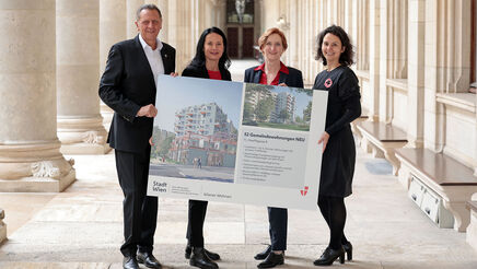 Drei weibliche und eine männliche Person stehen in den Arkaden des Wiener Rathauses und halten gemeinsam ein Schild auf dem Visualisierungen des Gemeindebau NEU zu sehen sind, sowie ein wenig Text