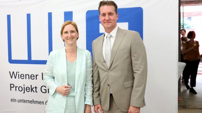 Zwei Personen in Anzügen stehen vor einem weißen Banner mit blauem Logo