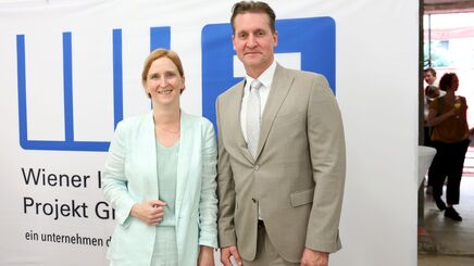 Zwei Personen in Anzügen stehen vor einem weißen Banner mit blauem Logo