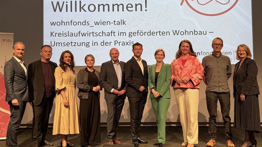 Gruppe von zehn Personen steht vor einer Präsentationswand mit dem Text 'Willkommen! wohnfonds_wien-talk Kreislaufwirtschaft im geförderten Wohnbau – Umsetzung in der Praxis'
