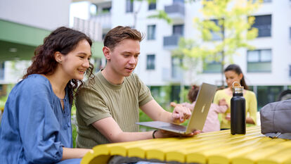 2 junge Menschen sitzen auf einer Parkbank vor einem Tisch und blicken erfreut auf den Laptop der vor ihnen steht. Im Hintergrund ist eine junge Frau zu erkennen