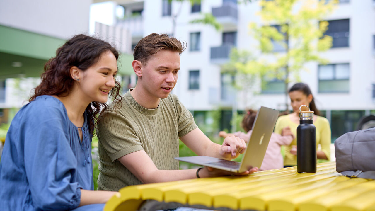 2 junge Menschen sitzen auf einer Parkbank vor einem Tisch und blicken erfreut auf den Laptop der vor ihnen steht. Im Hintergrund ist eine junge Frau zu erkennen