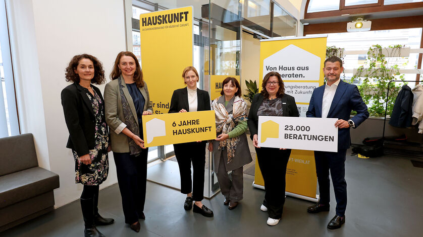 Fünf Personen stehen in einem modernen Innenraum und halten gelbe Schilder mit den Aufschriften '6 JAHRE HAUSKUNFT' und '23.000 BERATUNGEN', im Hintergrund gelbe Banner mit dem Schriftzug 'HAUSKUNFT'