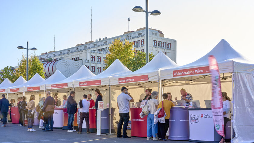 Menschen stehen an mehreren weißen Zelten mit roten Bannern und Informationsständen in einer sonnigen Straße vor einem großen Gebäude