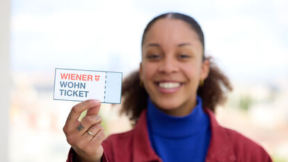 Imagebild, eine junge Frau hält ein Wiener Wohn-Ticket direkt in die Kamera, sie ist unscharf im Hintergrund, das Wohn-Ticket im Vordergrund