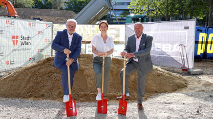 Drei Personen in formeller Kleidung stehen auf einer Baustelle und halten jeweils einen roten Spaten in der Hand, hinter ihnen ein großer Erdhaufen und Bauzäune mit dem Logo der Stadt Wien