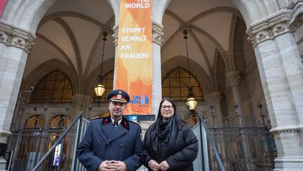 Ein hoher Polizeibeamter in Uniform mit Mantel und die Vizebürgermeisterin im Wintermantel posieren vor der am Wiener Rathaus gehissten orangen Flagge mit der Aufschrift "Orange the World" und "Stoppt Gewalt an Frauen!"