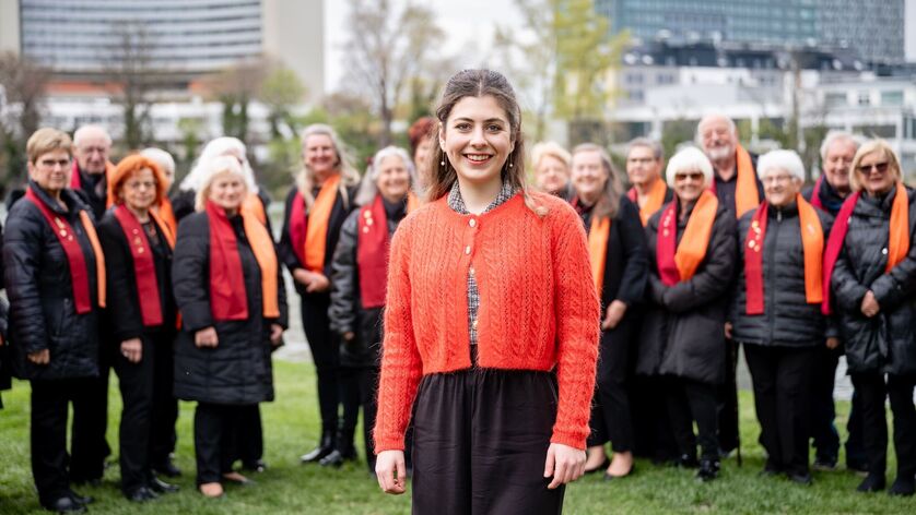 Frau mit rotem Strickjacke und schwarzer Hose steht im Vordergrund vor einer Gruppe von Personen mit schwarzen Outfits und orangen Schals auf einer Wiese in urbaner Umgebung