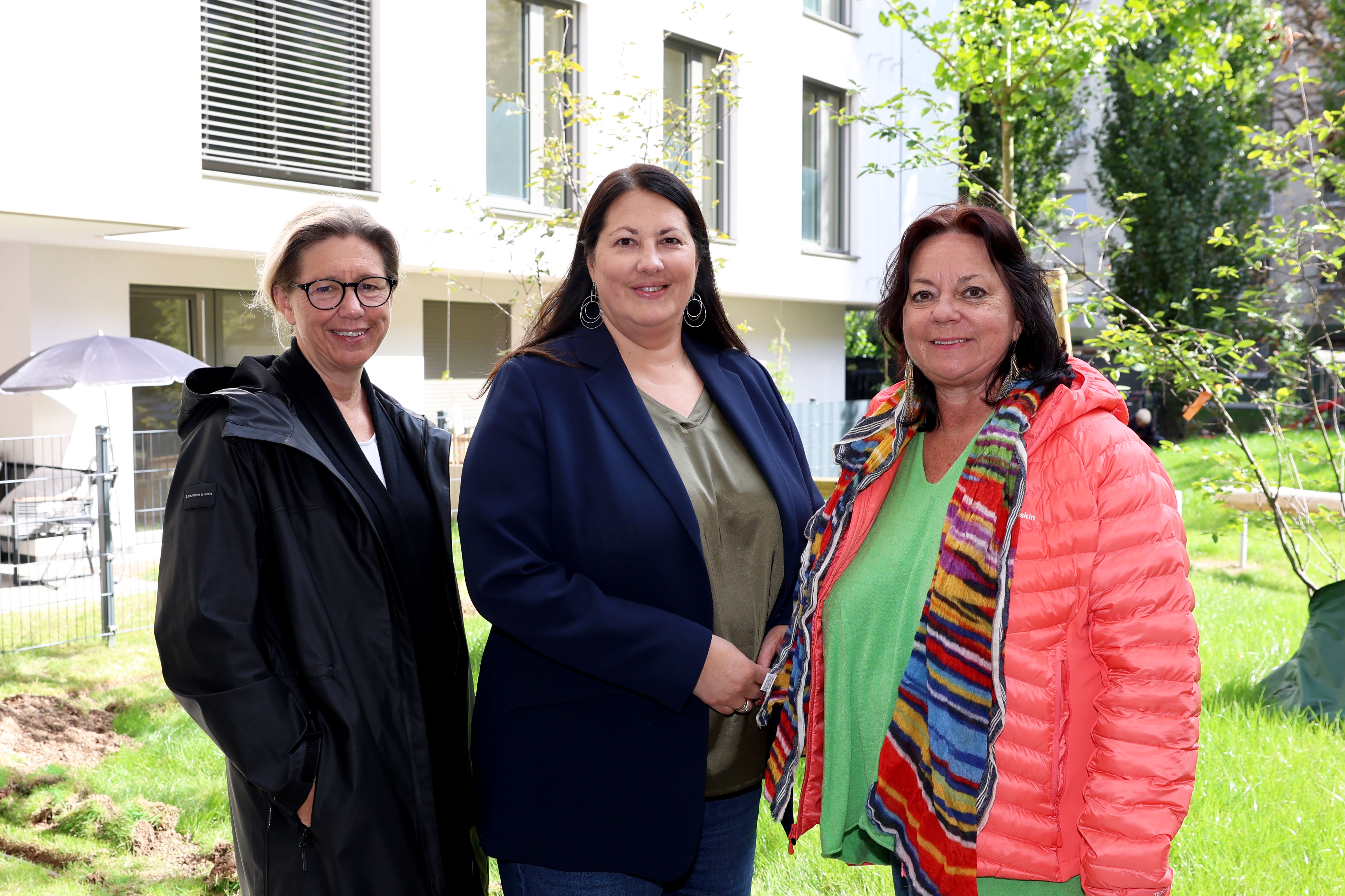Wiener Wohnen Direktorin Karin Ramser, Vizebürgermeisterin und Wohnbaustadträtin Kathrin Gaál und wohnpartner-Bereichsleiterin Claudia Huemer stehen im begrünten Innenhof des neuen Gemeindebaus. Die drei sind trotz des offensichtlich freundlichen Wetters herbstlich gekleidet und lächeln freundlich in die Kamera.